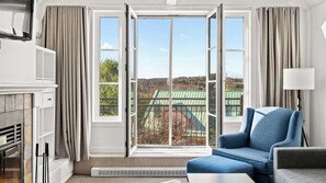 Ein Zimmer mit einer großen Schiebeglaswand, einem blauen Sessel, einem weißen Kamin und Blick auf ein grünes Gebäude.