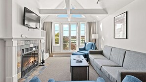Ein modernes Wohnzimmer mit Kamin, Fernseher, Sofa, Sessel und großen Fenstern mit Blick auf Grünflächen.