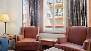 Ein Zimmer mit zwei Sesseln, einem kleinen Tisch, einer Lampe und einem Fenster mit Blick auf ein Gebäude.
