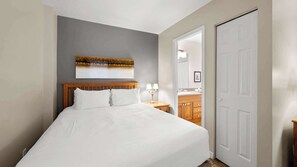 Ein ordentlich bezogenes Bett in einem Hotelzimmer, mit einem hölzernen Kopfteil, einem Nachttisch mit Lampe, einem Spiegel und einer Tür zu einem Badezimmer.