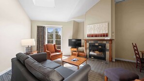 Ein Wohnzimmer mit einer Couch, einem Sessel, einem Couchtisch, einem Kamin, einem Fernseher und einem fenster mit Vorhängen.