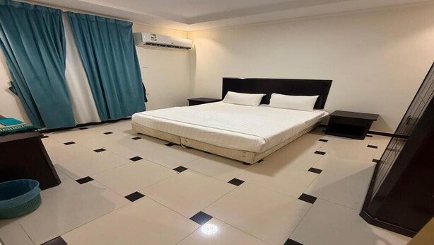 A hotel room with a bed, a nightstand, a desk, a chair, a blue trash bin, and a window with curtains. - Quiet Rooms 13 (Riyadh)