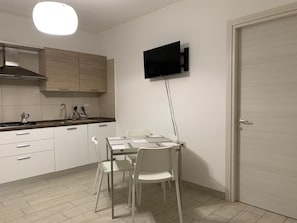 Une cuisine moderne avec des meubles blancs, un téléviseur à écran plat fixé au mur, une table à manger avec des chaises et une porte sur le côté droit.