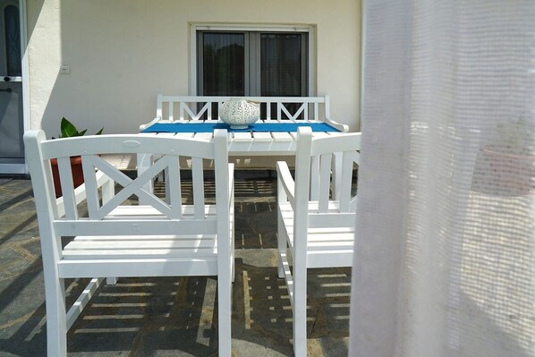 Un espacio exterior con mobiliario blanco, una mesa, sillas, una planta en maceta y una ventana con cortinas.