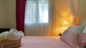 A hotel room with a bed, a bedside lamp, a window with sheer curtains, and a wicker basket with towels. - Apartment '1' with Mountain View, Wi-Fi and Air Conditioning (Fanari)