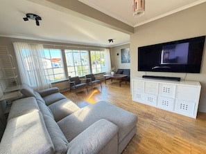 A spacious living room with a large grey sectional sofa, a wooden coffee table, a TV on a white cabinet, and large windows with blinds. - Family friendly house centrally located in Reykjavik (Reykjavík)