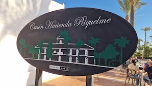 En skylt för Casón Hacienda Riquelme med en avbildning av en byggnad och palmer.