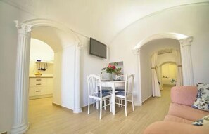 A dining area with a table and chairs, a television above the table, and a kitchen in the background. - Los Mosaicos Cave. Sleep in a Luxury Cave House with a Sunny Terrace—Guadix (Guadix)