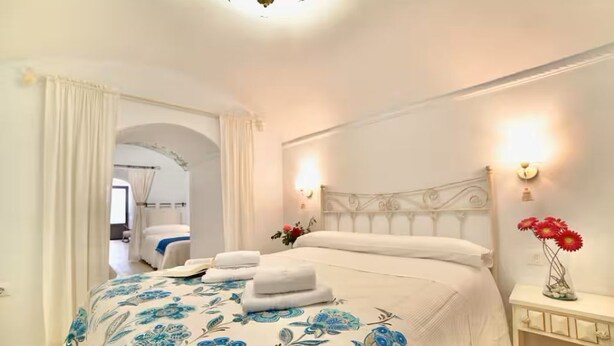 Een slaapkamer met een bed, witte gordijnen, een nachtkastje met een bloevas, en een wandlamp.