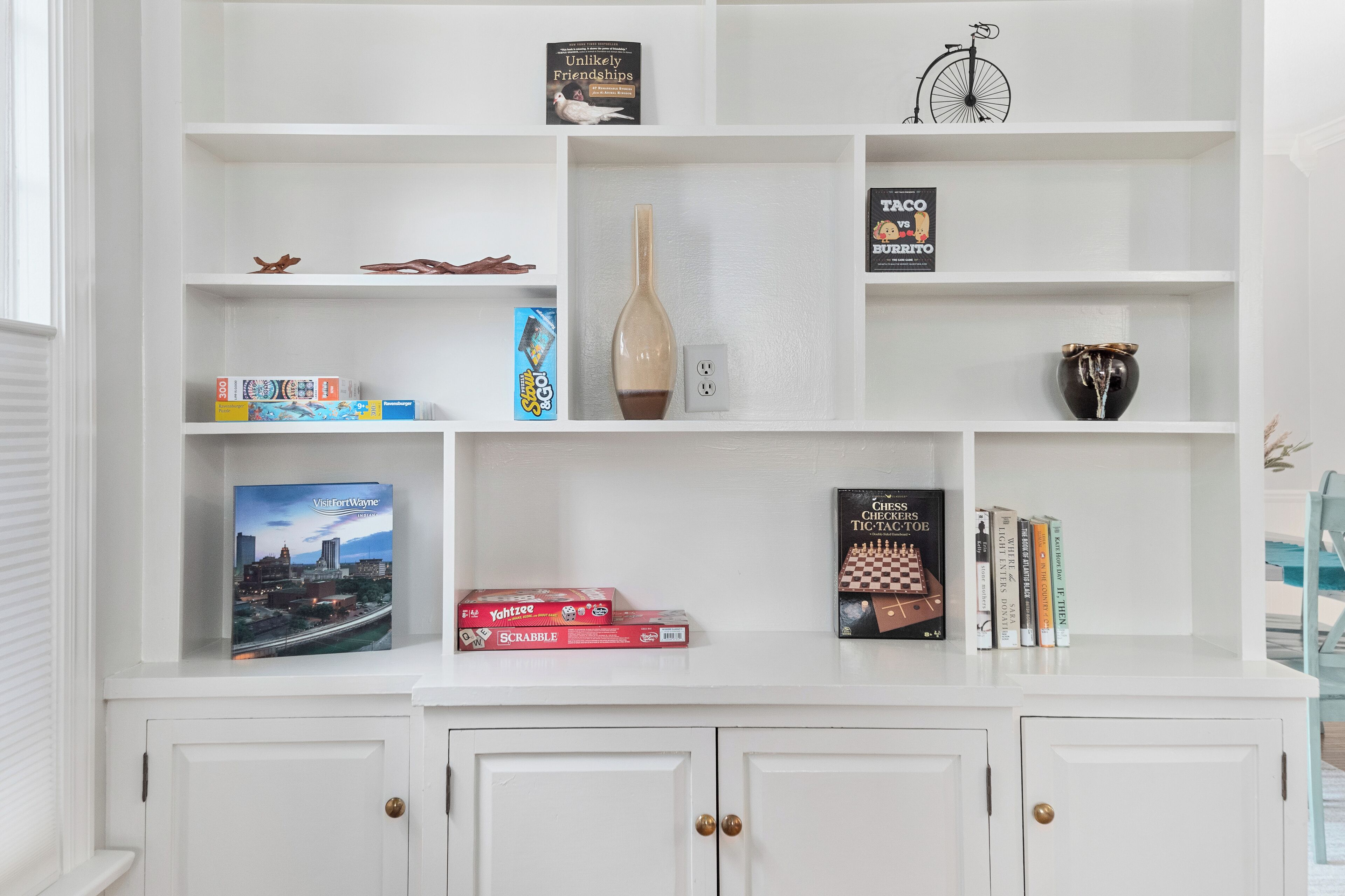 A white bookshelf with various board games, a vase, and a book titled 'Unlikely Friendships'.