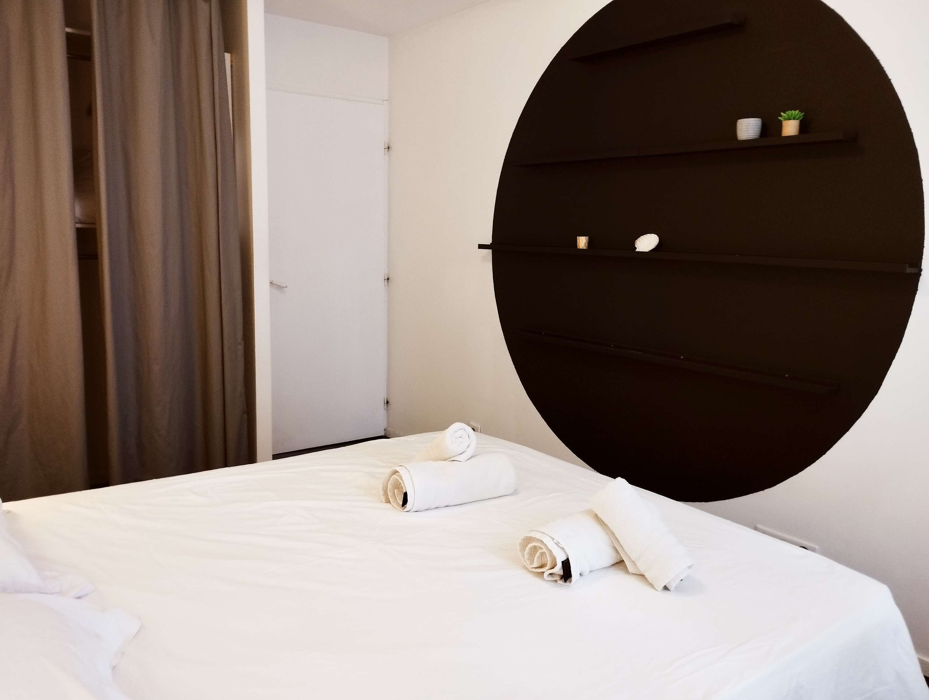 Une chambre d’hôtel avec un lit, une étagère circulaire fixée au mur et ornée d’une petite plante, et une porte.