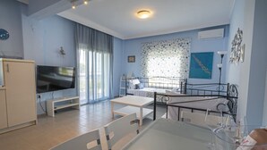 A room with a TV, a dining table with chairs, a bed, a window with curtains, and a wall with framed pictures. - Sandra Seafront Apartment by RentalsPro - Moudania Halkidiki (Nea Moudania)