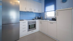 A kitchen with white cabinets, a stainless steel refrigerator, a built-in oven, and a window with a clock. - Sandra Seafront Apartment by RentalsPro - Moudania Halkidiki (Nea Moudania)