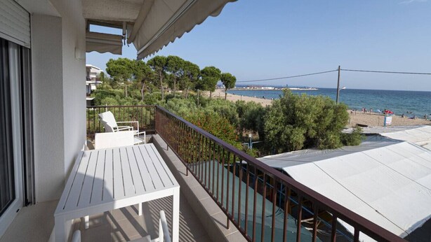 A balcony with a white table and chairs, overlooking a beach with trees and a clear sky. - Sandra Seafront Apartment by RentalsPro - Moudania Halkidiki (Nea Moudania)