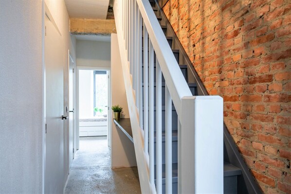 Un pasillo con una escalera blanca, un muro de ladrillo y una puerta que da a otra habitación.