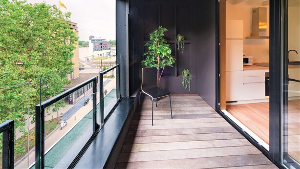 Un balcon avec un plancher en bois, une chaise, un pot de fleurs et une vue sur une rue et des immeubles.