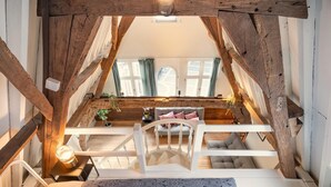 A loft bedroom with a wooden staircase, a bed with patterned bedding, a small table with a lamp, and a view of a living room with a sofa and plants. - Loft-style Duplex at Hendrik Square (Antwerp)