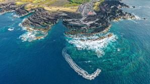 Vista aerea di una zona costiera con formazioni rocciose, acque blu cristalline e una strada che conduce a un edificio con tetto rosso.