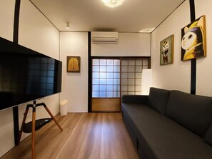 A modern living room with a large flat-screen TV on a tripod stand, a dark sofa, a tatami mat, and a shoji screen door. - Leisureyado/5minTrain to USJ/2BR PrivateHouse/Direct to KIX (Osaka)