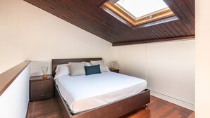 Una camera da letto con finestra a lucernario, soffitto in legno, un letto con biancheria da letto bianca, un comodino e un mobiletto contenitore.