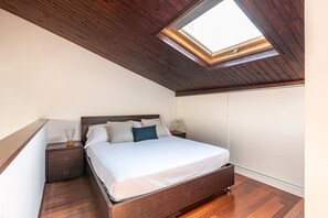 Quarto com claraboia, teto de madeira, cama com roupa de cama branca, mesa de cabeceira e uma cômoda.