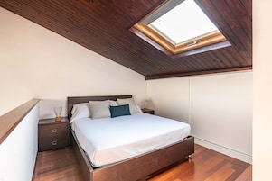 Un dormitorio con tragaluz, techo de madera, cama con ropa de cama blanca, mesita de noche y cómoda.