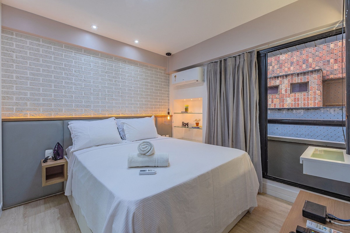 Une chambre d’hôtel avec un mur décoré d’un motif en briques, un lit avec du linge de lit blanc, une table de chevet avec une lampe et une fenêtre donnant sur un immeuble.