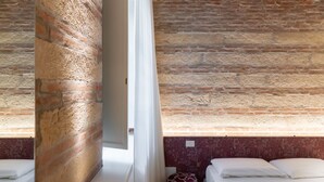 Een kamer met onbehandelde bakstenen muren, een bed met wit beddengoed en een gestreept voetenbankje.
