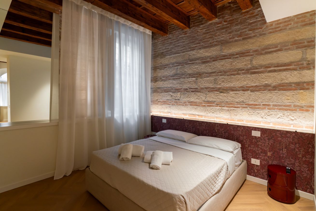 A bedroom with a wooden ceiling, a brick wall, a bed with white linens, and a red trash bin.