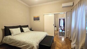 Quarto com cama, um banco e porta que dá para outro cômodo.