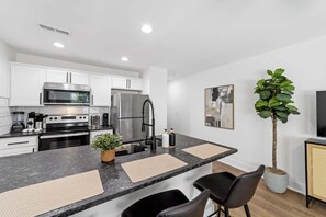 A modern kitchen with white cabinets, stainless steel appliances, a central island with two chairs, and a potted plant. - 1BD Modern Condo ⎮ Downtown Walkable to F&M Bank Arena (Clarksville)