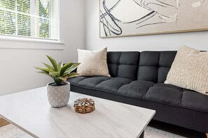 A modern living room with a dark sofa, a marble coffee table, a potted plant, a woven basket, and a large abstract painting on the wall. - 1BD Modern Condo ⎮ Downtown Walkable to F&M Bank Arena (Clarksville)