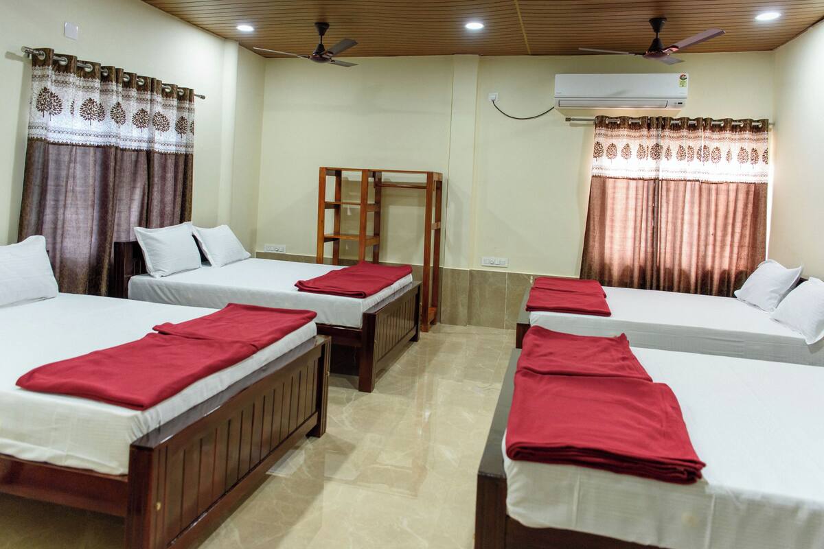 A hotel room with four single beds, each with a red towel, wooden headboards, and white bedding. There is a ceiling fan, an air conditioning unit, and a wooden chair in the center.