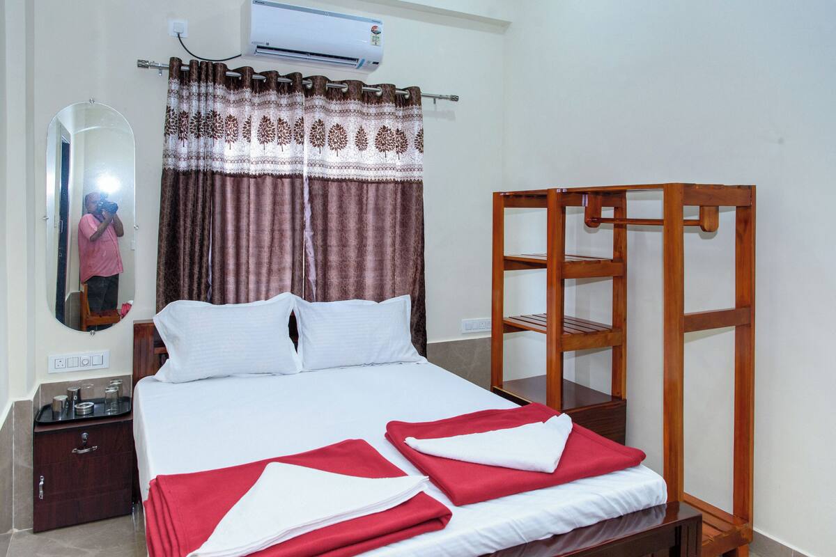 A hotel room with a bed, a wooden side table, a mirror, and a window with curtains.