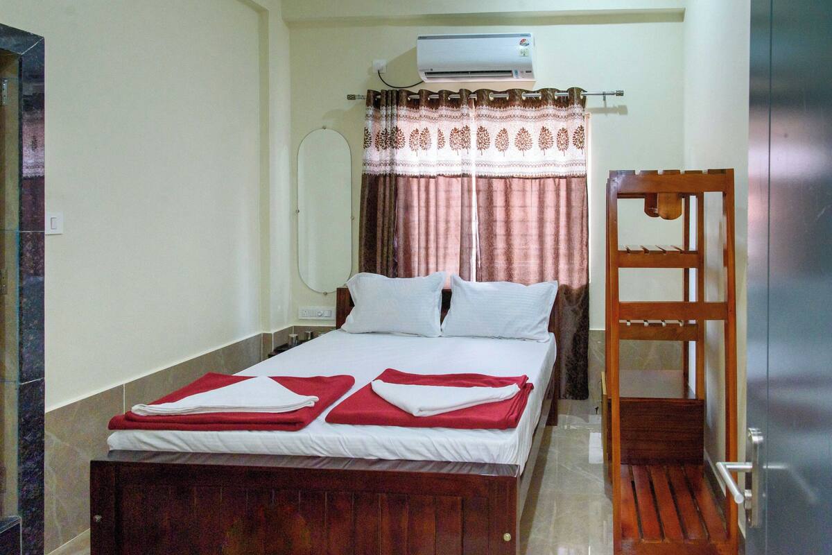 A hotel room with a bed, a wooden chair, an air conditioning unit, and a curtain.