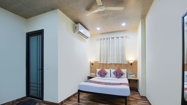 A bedroom with a bed, a ceiling fan, an air conditioning unit, a nightstand, and a window with curtains. - Hotel Royal Residency (New Delhi)