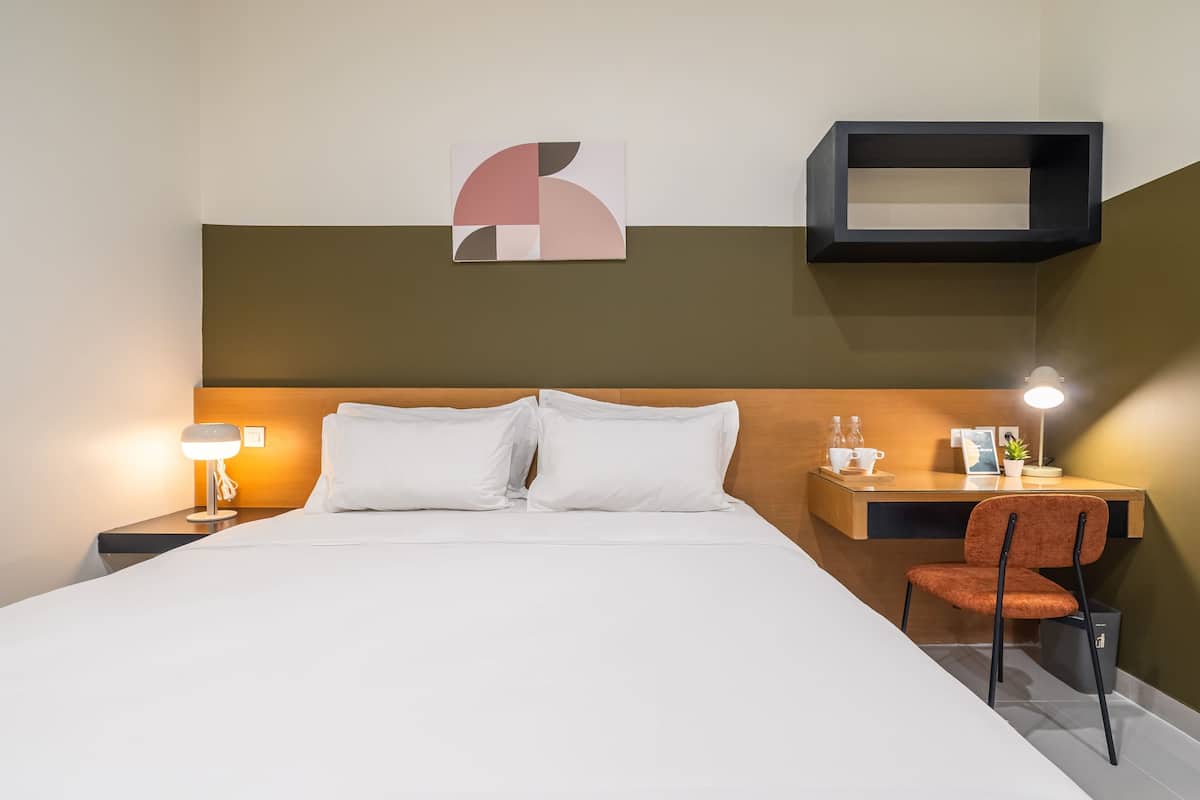 Habitación de hotel con cama, escritorio, silla, mesita de noche, lámpara, una obra de arte en la pared y estante.