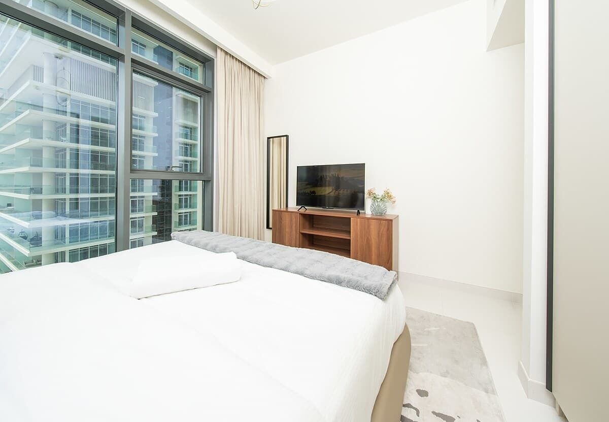 A modern hotel room with a large bed, a TV, and a view of a building through a large window.