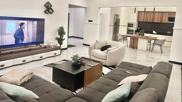 A modern living room with a flat-screen TV, sofas, a coffee table, and a kitchen area in the background.