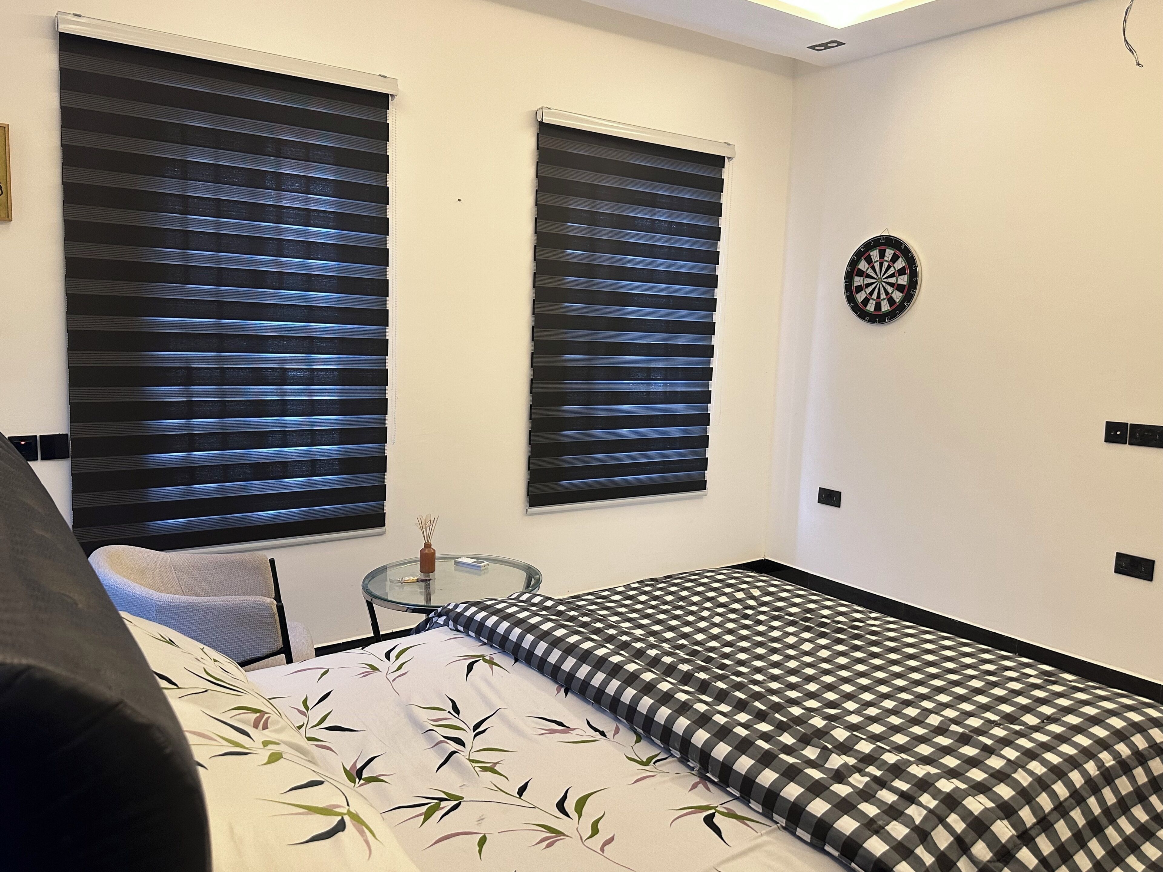 A bedroom with a bed, two windows with blinds, a small round table, and a dartboard on the wall.
