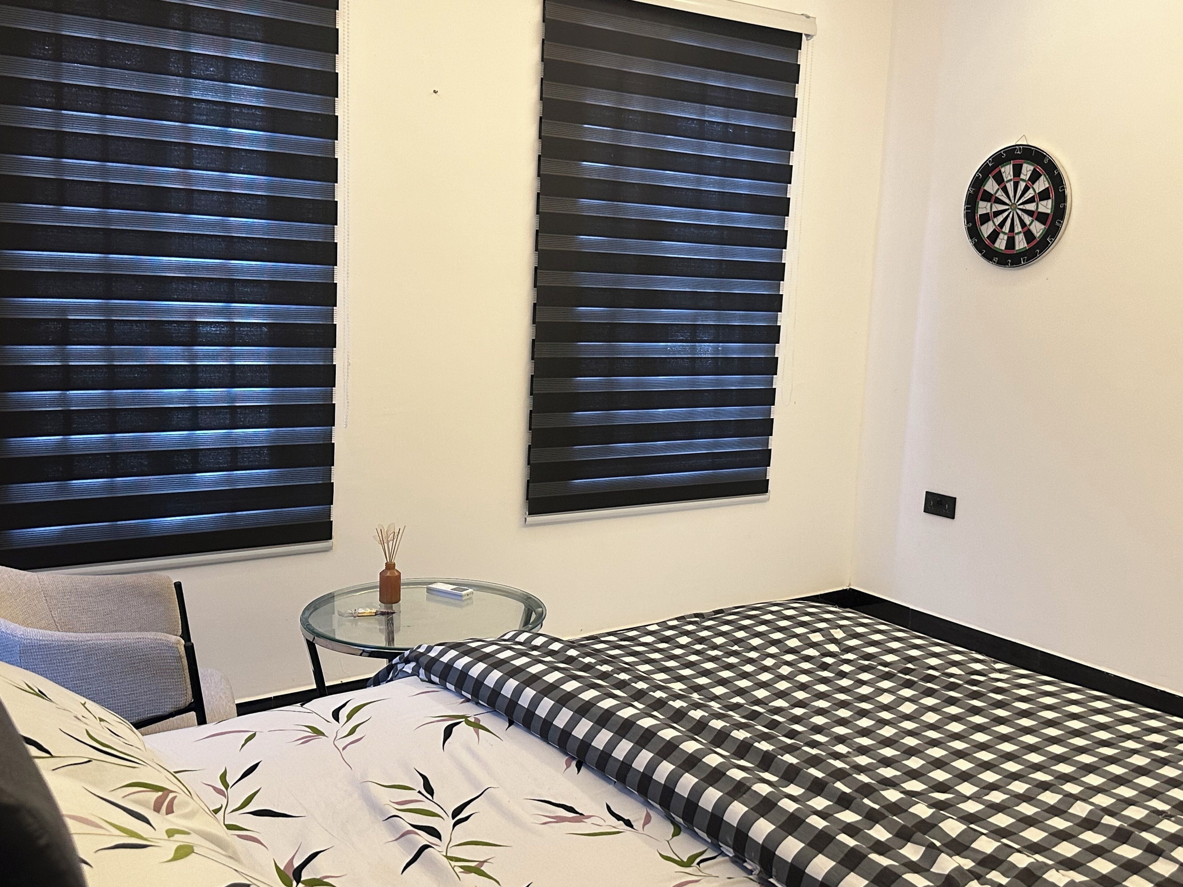 A room with a bed, a glass table with a vase, and two windows with blinds.