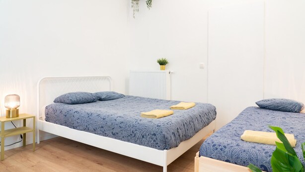 Ein Zimmer mit zwei Betten, einem Nachttisch mit Lampe, einem Heizkörper und einer kleinen Pflanze.