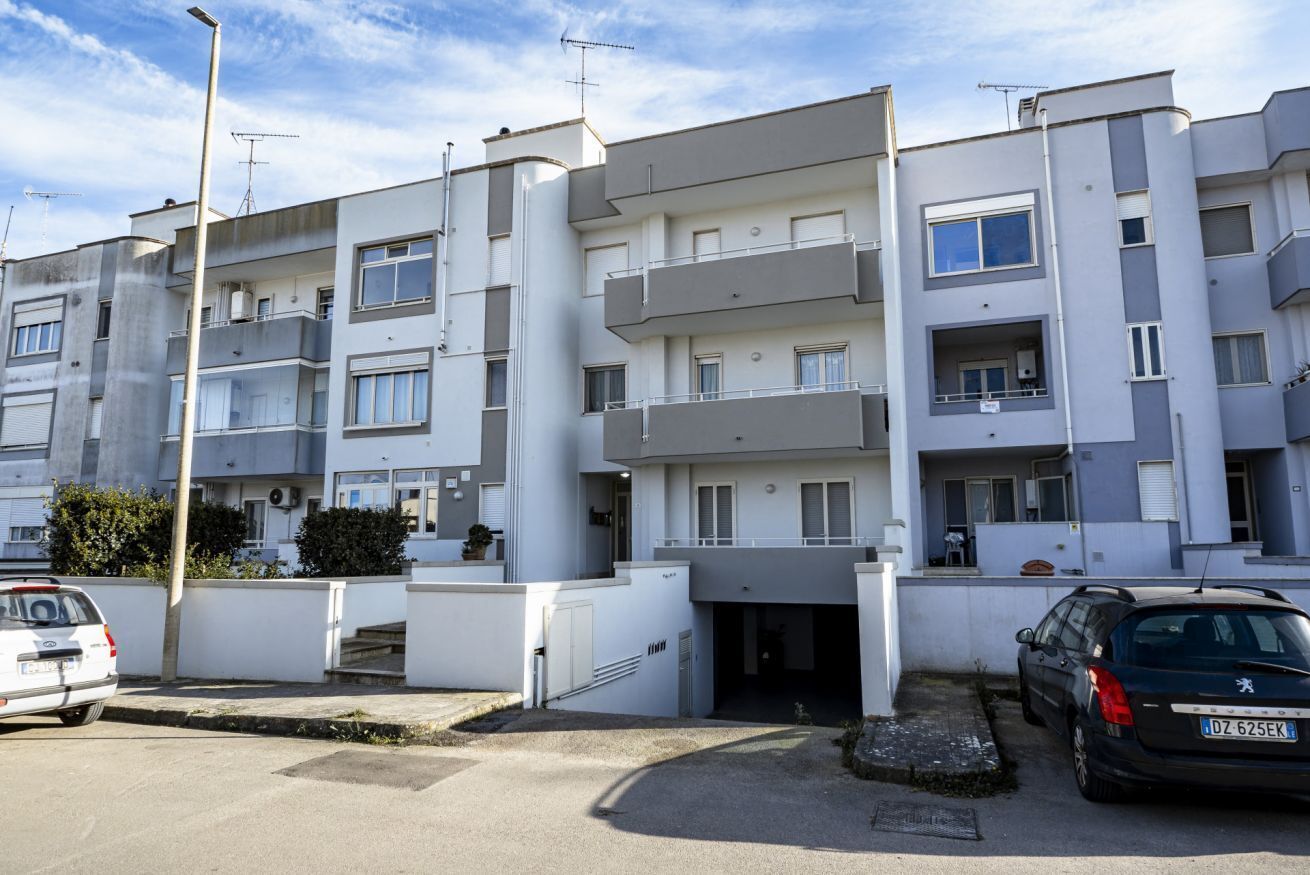 A multi-story residential building with balconies, a parking area, and a clear sky.