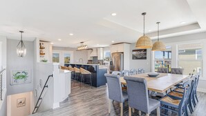 A modern kitchen with a central island, a dining area with a wooden table and chairs, and a staircase leading to another level. - The Crooked Palm | Luxury Coastal Escape w/ Pool & Views | Steps to the Beach (Oak Island)