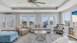 A spacious living room with a ceiling fan, a sofa set, a coffee table, and a view of the beach through large windows. - The Crooked Palm | Luxury Coastal Escape w/ Pool & Views | Steps to the Beach (Oak Island)