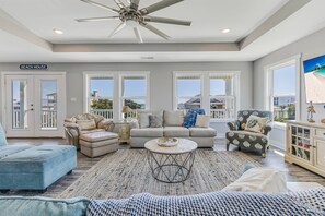 A spacious living room with a ceiling fan, a sofa set, a coffee table, and a view of the beach through large windows. - The Crooked Palm | Luxury Coastal Escape w/ Pool & Views | Steps to the Beach (Oak Island)