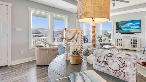 A living room with a round table, a lamp, a decorative sign, and a television. - The Crooked Palm | Luxury Coastal Escape w/ Pool & Views | Steps to the Beach (Oak Island)