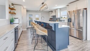 A modern kitchen with a central island, bar stools, stainless steel appliances, and a view to the outside. - The Crooked Palm | Luxury Coastal Escape w/ Pool & Views | Steps to the Beach (Oak Island)