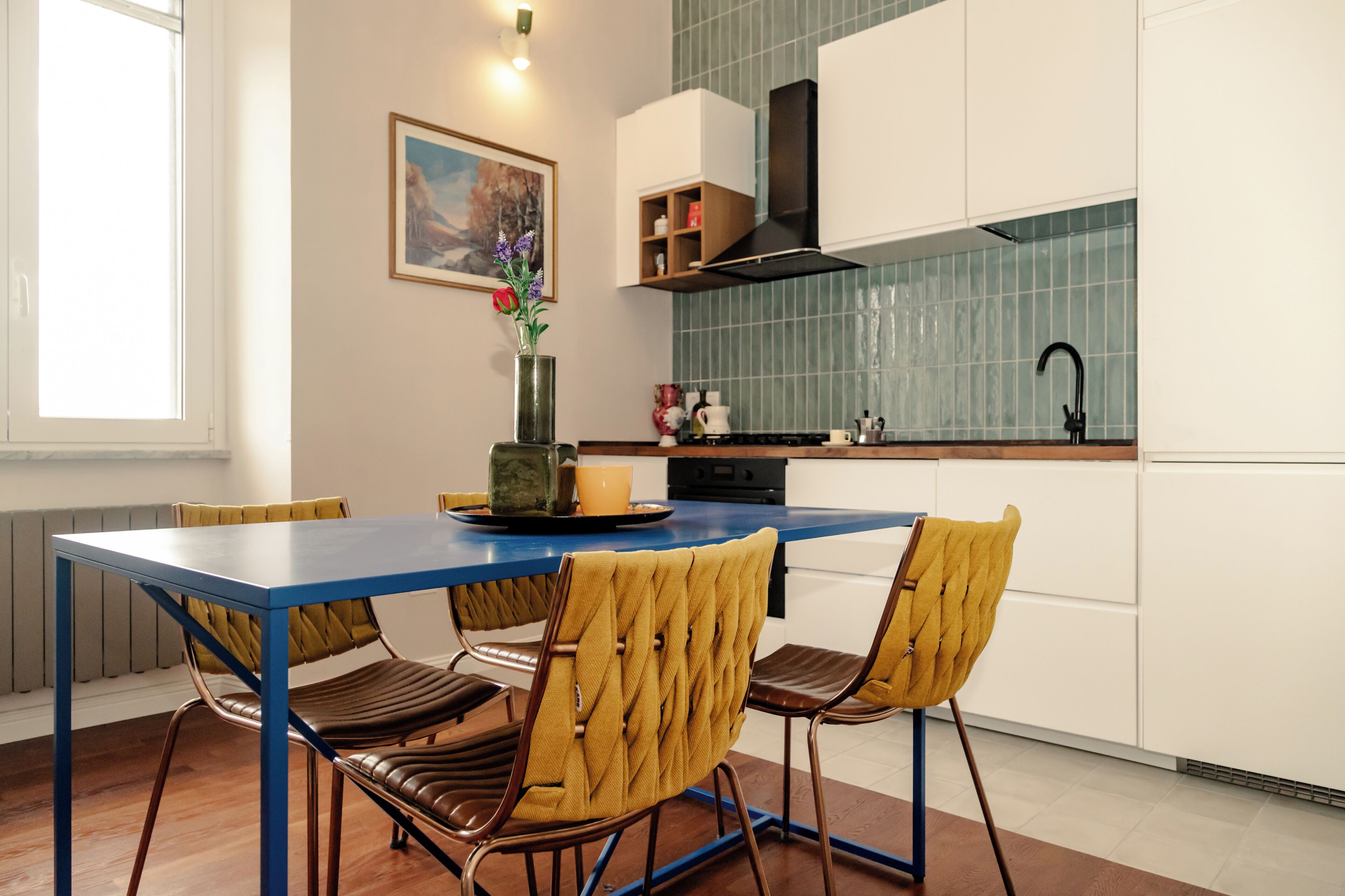 Une cuisine moderne avec une table bleue, deux chaises jaunes, un vase de fleurs et un tableau accroché au mur.