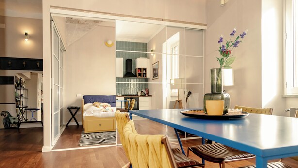 A modern living room with a blue dining table, yellow chairs, a sofa, and a kitchen in the background. - Loft Pop-Industrial con vista Acquedotto (Rome)